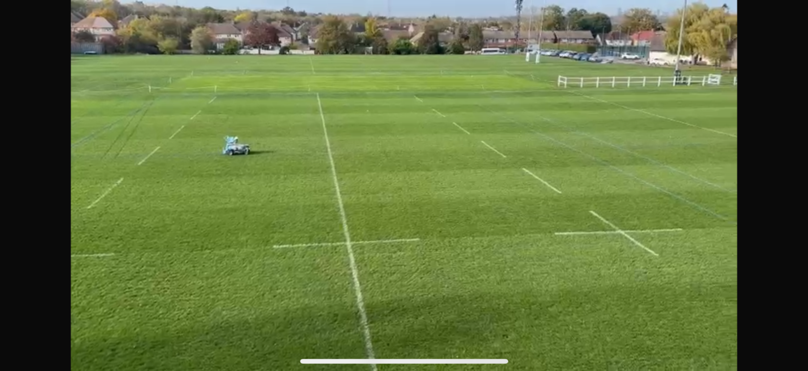 A wide view of a green rugby pitch with marked lines and a player on the field.